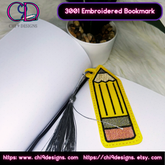 An embroidered pencil bookmark with a silver tassel placed inside an open book. The bright yellow design adds a playful touch, perfect for marking your page in style