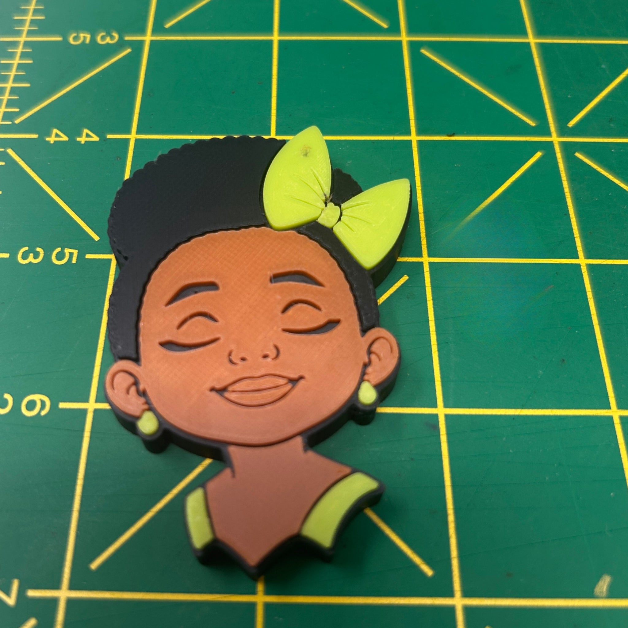 A 3D-printed cartoon-style female character head charm or plaque, featuring a black hair bun with a lime green bow, closed eyes, and a green top, placed on a green cutting mat with yellow grid lines.