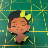 A 3D-printed cartoon-style female character head charm or plaque, featuring a black hair bun with a lime green bow, closed eyes, and a green top, placed on a green cutting mat with yellow grid lines.