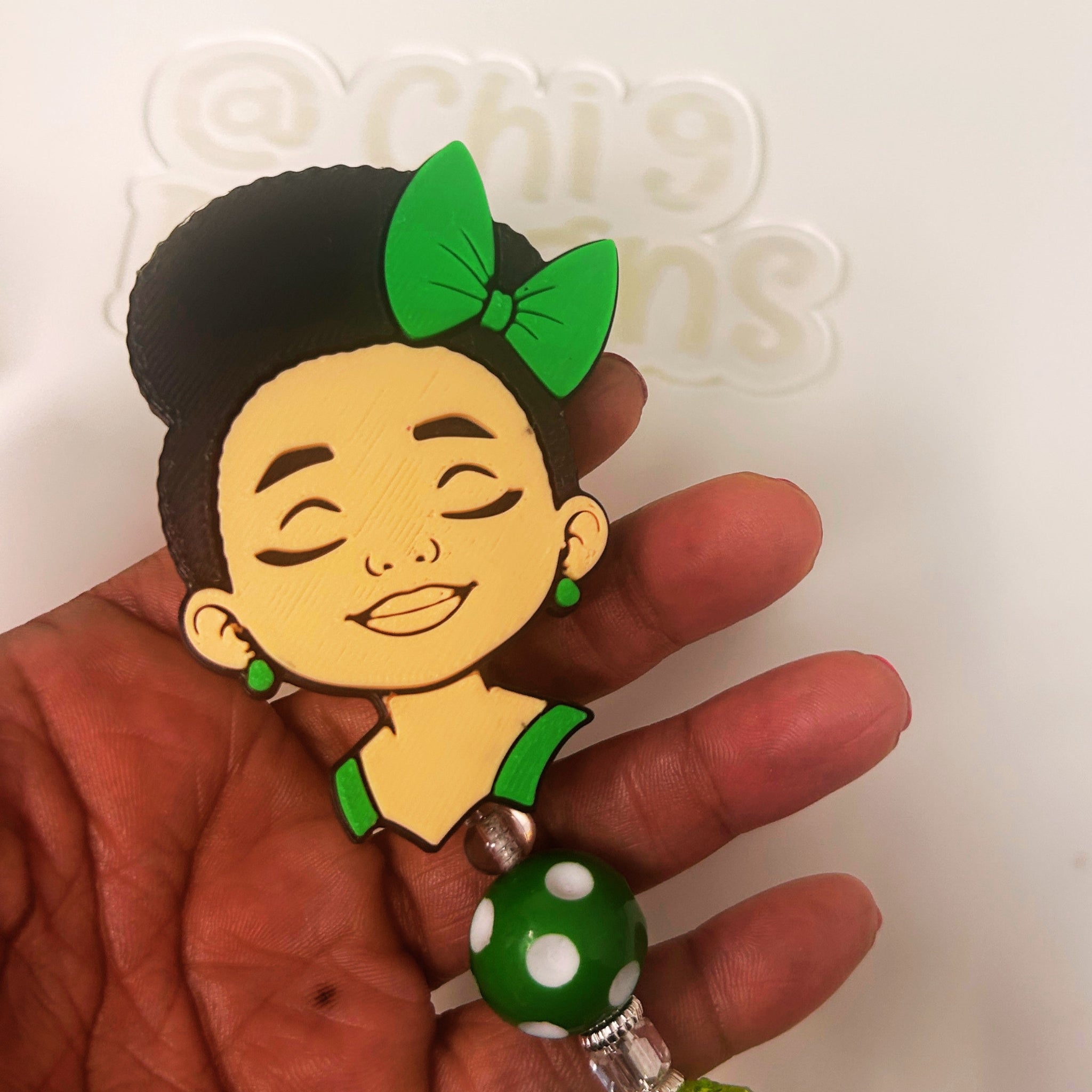 A hand holding a cartoon-style badge reel featuring a woman with black hair, a green bow, and green earrings, with a green and white polka dot bead hanging below.