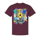 A maroon short-sleeve crewneck t-shirt featuring a colorful 'FIFTH grade' graphic on the front, complete with a lightbulb, pencil, notebook, and stars.