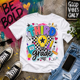 including black sneakers, sunglasses, a phone, ripped blue jeans, and paint splatter art. The shirt features a colorful retro-style 'THIRD grade' graphic with a lightbulb, stars, hearts, and doodles.