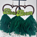 A pair of dangling earrings with dark brown heart-shaped cutouts at the top, bright green script text reading 'Bloom Bloom' across the center, and large dark green yarn fringe below, displayed on a black stand against a white background.
