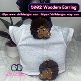 A pair of Afrocentric wooden earrings featuring a bold woman's face with an Afro hairstyle, accented with fabric inlay, glitter, and decorative stones, displayed on a mannequin wearing a white top.