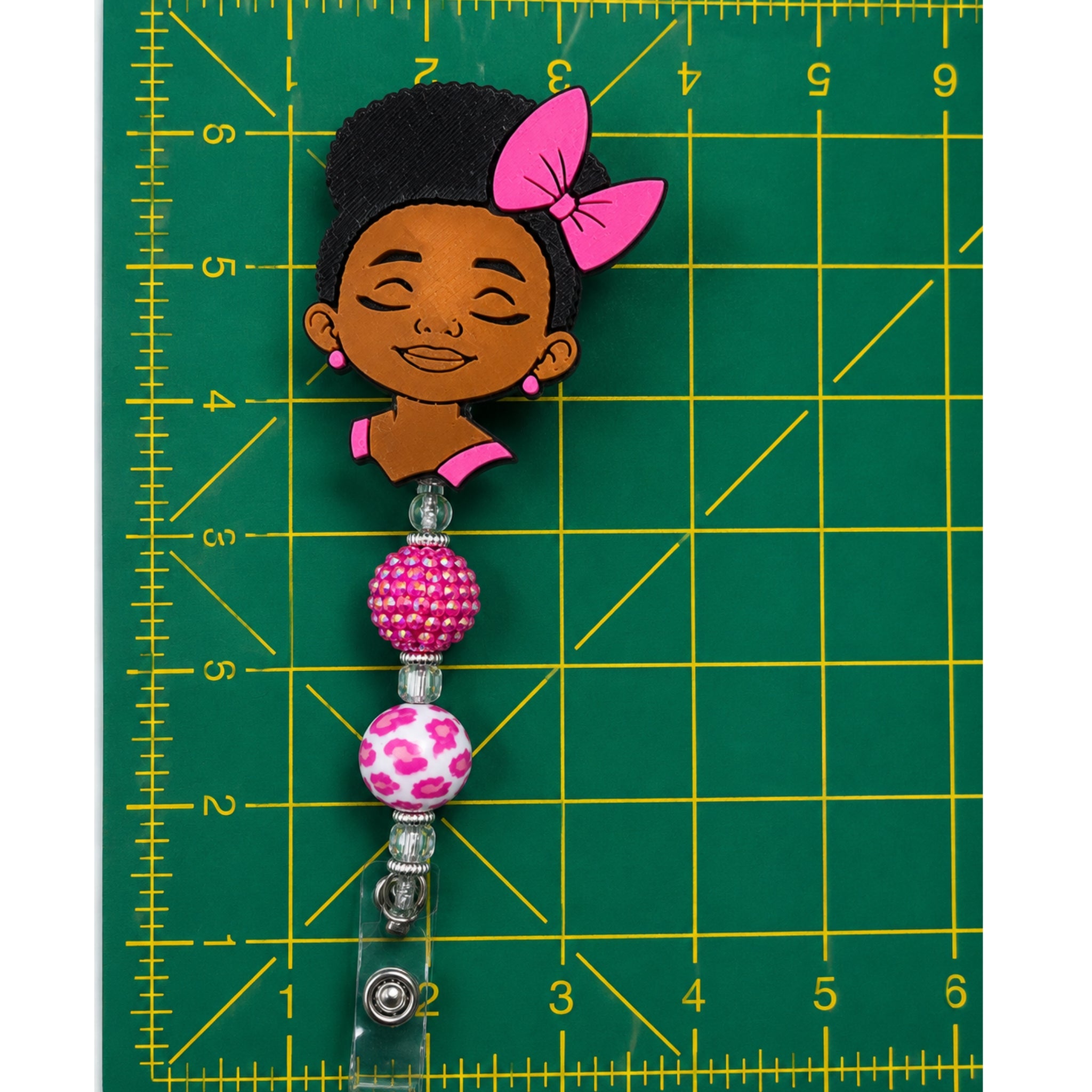A handmade beaded badge reel pin clasp featuring a cartoon girl charm with black hair and a pink bow, attached to pink and white patterned beads, shown on a green cutting mat with yellow grid lines.