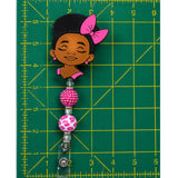 A handmade beaded badge reel pin clasp featuring a cartoon girl charm with black hair and a pink bow, attached to pink and white patterned beads, shown on a green cutting mat with yellow grid lines.