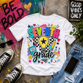 A white graphic T-shirt laid flat on a concrete surface with styled props including black sneakers, sunglasses, a phone, jeans, and paint splatter decor. The shirt features a colorful 'SEVENTH grade' graphic with a lightbulb, stars, hearts, and school-themed doodles.