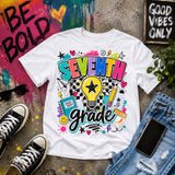 A white graphic T-shirt laid flat on a concrete surface with styled props including black sneakers, sunglasses, a phone, jeans, and paint splatter decor. The shirt features a colorful 'SEVENTH grade' graphic with a lightbulb, stars, hearts, and school-themed doodles.