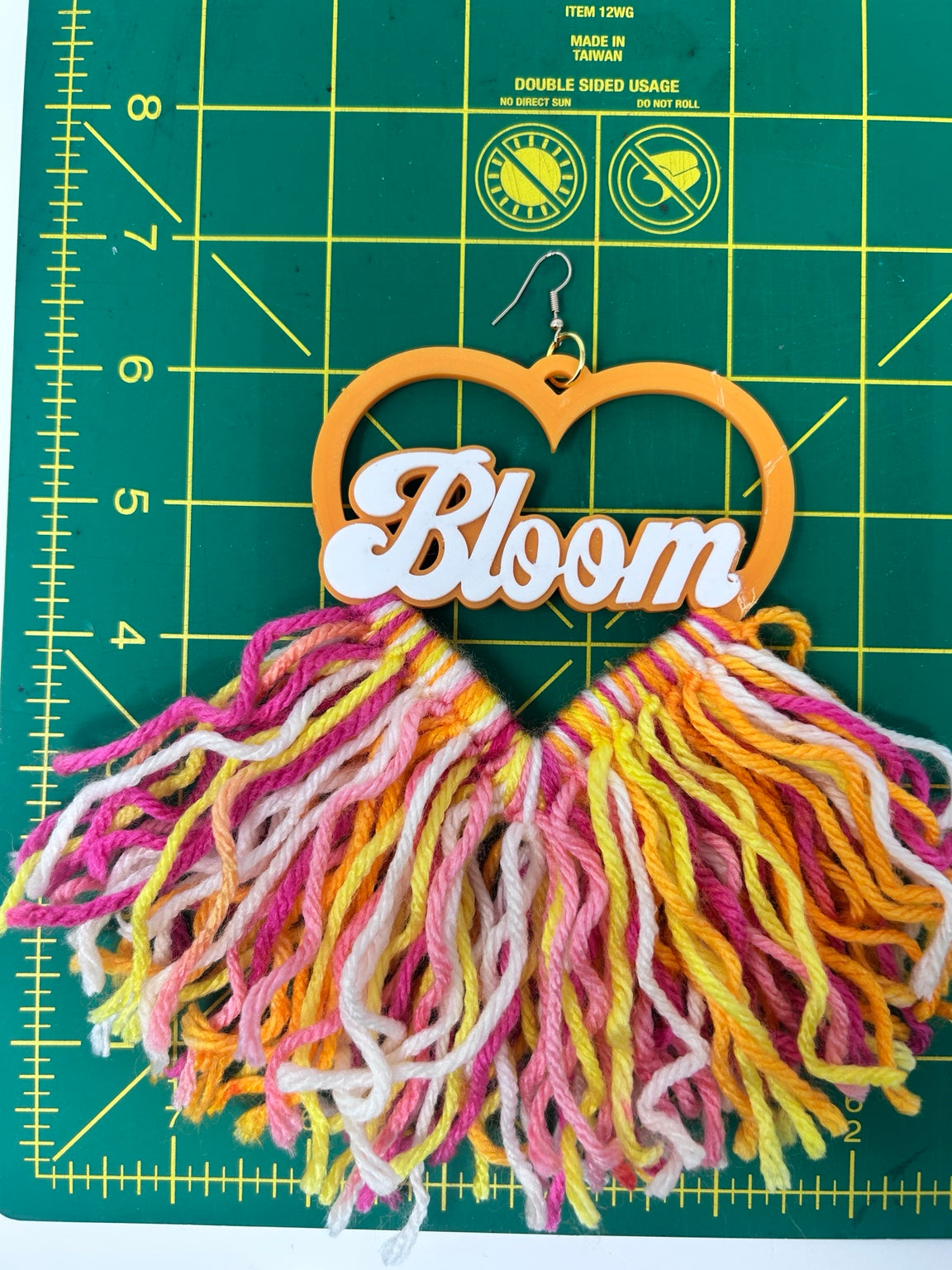 A handmade wooden earring featuring a heart-shaped top with the word 'Bloom' in white script, hanging from a silver fishhook. The bottom is a large, fluffy tassel made of multi-colored yarn in pink, yellow, orange, and white.