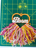 A handmade wooden earring featuring a heart-shaped top with the word 'Bloom' in white script, hanging from a silver fishhook. The bottom is a large, fluffy tassel made of multi-colored yarn in pink, yellow, orange, and white.