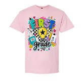 A light pink short-sleeve t-shirt with a colorful 'FIRST grade' graphic on the front, featuring a light bulb, pencils, notebook, and stars.