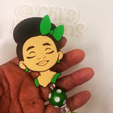 A hand holding a cartoon-style badge reel featuring a woman with black hair, a green bow, and green earrings, with a green and white polka dot bead hanging below.