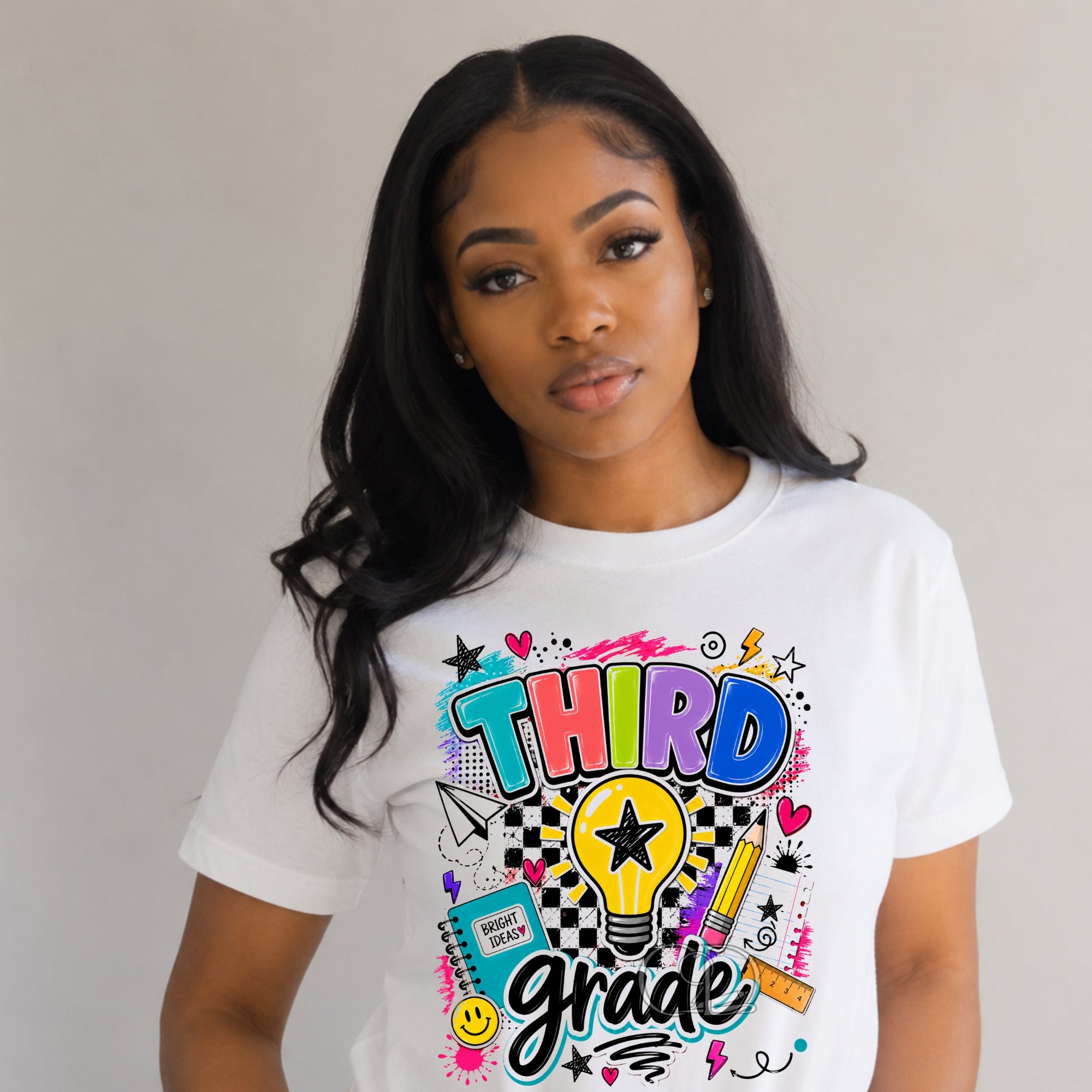 A woman wearing a white short-sleeve graphic T-shirt with a colorful 'THIRD grade' design featuring a lightbulb, stars, hearts, and school-themed doodles, set against a plain light gray background.