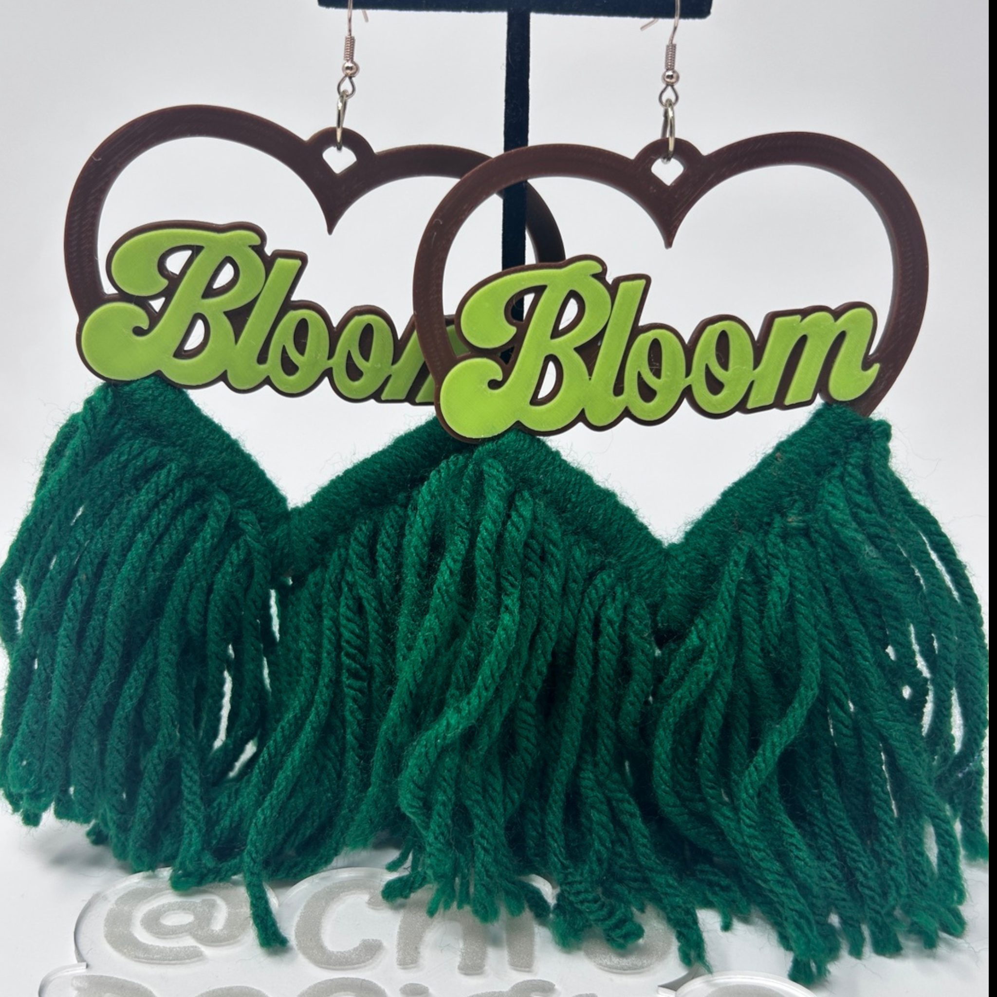 A pair of dangling earrings with dark brown heart-shaped cutouts at the top, bright green script text reading 'Bloom Bloom' across the center, and large dark green yarn fringe below, displayed on a black stand against a white background.