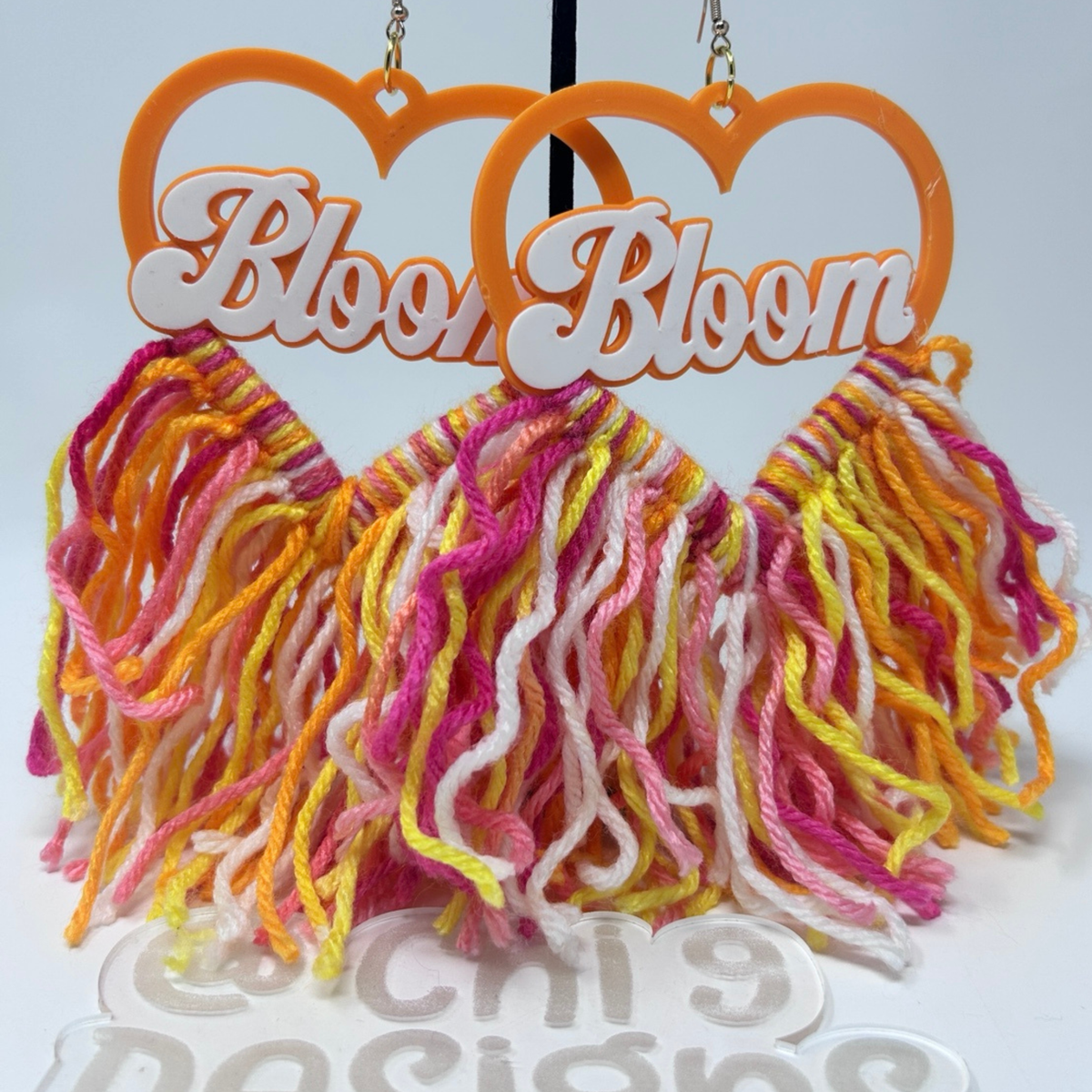 A pair of dangling earrings with bright orange heart-shaped tops featuring the word 'Bloom' in white script, and multiple strands of variegated yarn tassels in pink, orange, yellow, and white hanging below.