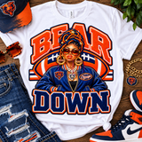 A white graphic T-shirt laid flat on a wooden surface, featuring a Chicago Bears-themed design with the words 'BEAR DOWN' and an illustrated woman in a blue jacket and headwrap. Surrounding props include a Chicago Bears cap, sunglasses, distressed jeans, a gold chain, a Bears patch, and sneakers.