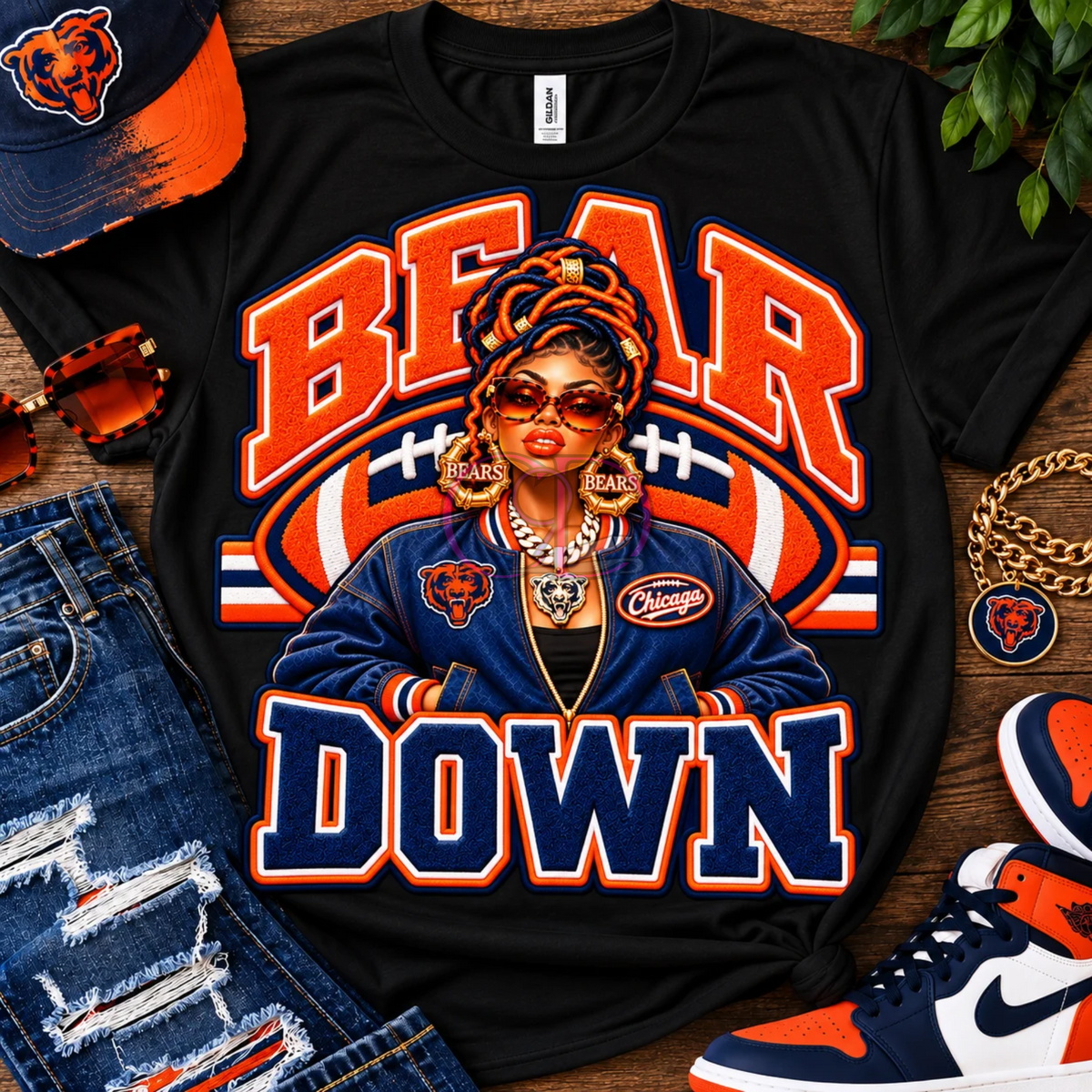 A black graphic T-shirt laid flat on a wooden surface, featuring a stylized Chicago Bears-themed illustration with the words 'BEAR DOWN' in orange and navy. The shirt is styled with a matching cap, sunglasses, distressed jeans, a gold chain, and sneakers.