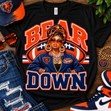 A black graphic T-shirt laid flat on a wooden surface, featuring a stylized Chicago Bears-themed illustration with the words 'BEAR DOWN' in orange and navy. The shirt is styled with a matching cap, sunglasses, distressed jeans, a gold chain, and sneakers.
