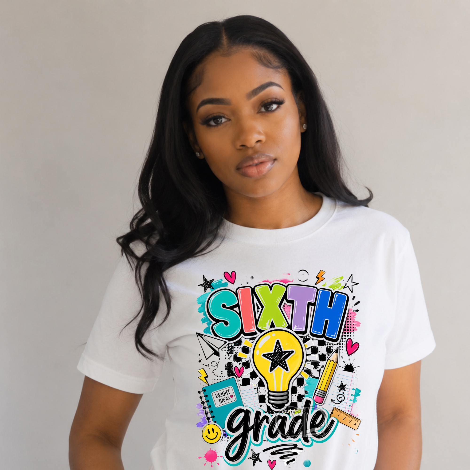 A woman wearing a white short-sleeve graphic T-shirt with colorful school-themed text reading 'SIXTH grade' and doodles, photographed against a plain light gray background.